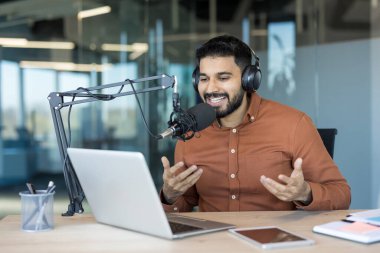 Modern bir ofisten canlı yayına ev sahipliği yapan genç adam, profesyonel kulaklıkları, laptopu açık, gülümseyen ve kendinden emin bir şekilde mikrofona konuşuyor.