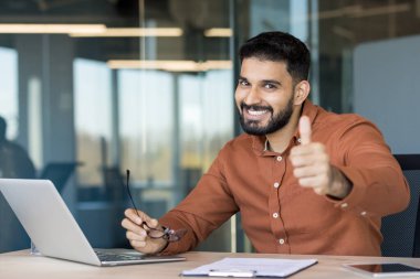 Modern bir ofisteki gülümseyen Hintli işadamı, üretken uzaktan çalışma ve liderlik süresince mesleki başarı, güven ve memnuniyeti onaylıyor.