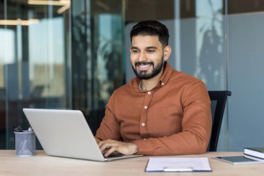 Kendine güveni tam genç Hintli bir adam dizüstü bilgisayar kullanıyor ve çağdaş bir şirkette masa başında çalışırken gülümsüyor, üretkenliğini ve dijital iletişimini gösteriyor.