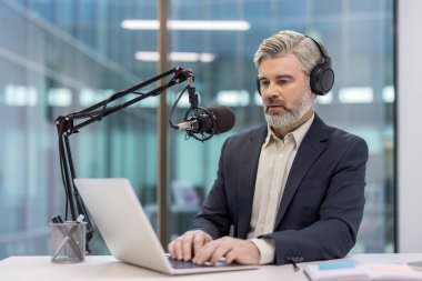 Kulaklık takan, kulaklık takan, stüdyo mikrofonuna konuşan, modern bir ofis masasında dizüstü bilgisayar kullanan, profesyonel uzaktan kumandalı yetişkin bir adam.