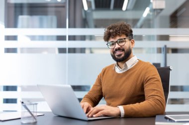 Sakallı ve gözlüklü genç adam kameraya gülümsüyor, masada oturuyor ve çağdaş ofis ortamında dizüstü bilgisayar kullanıyor, profesyonelliği ve başarıyı somutlaştırıyor.