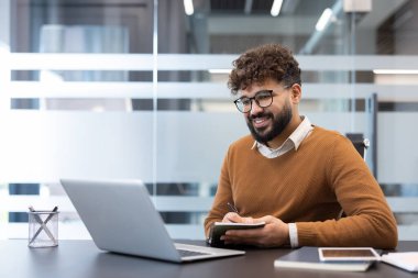 Genç adam gülümsüyor ve modern bir ofis masasında dizüstü bilgisayarla çalışırken not alıyor, üretkenlik ve profesyonelliğe odaklanıyor.