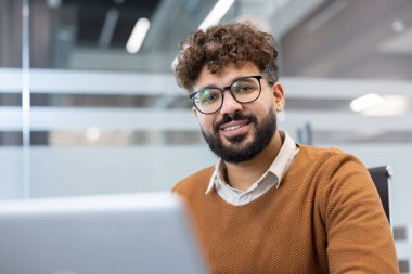 Sakallı, gözlüklü, kameraya gülümseyen, elinde dizüstü bilgisayarla masada oturan, iş başarısını ve profesyonel başarısını temsil eden kendine güvenen genç bir adam.