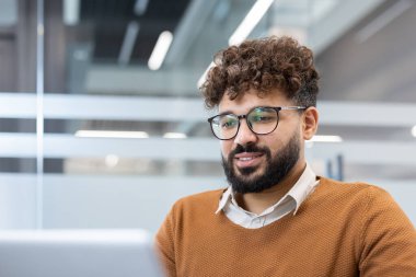 Çağdaş ofis ortamında çalışırken dizüstü bilgisayarına odaklanan kıvırcık saçlı ve gözlüklü genç sakallı adam özveri ve konsantrasyon sergiliyor.
