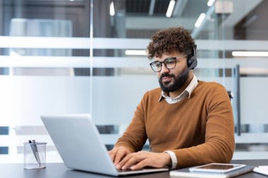Kıvırcık saçlı, gözlük takan ve kulaklık takan genç sakallı bir adam dizüstü bilgisayarda masa başında çalışıyor. Modern ofis ortamında online müşteri hizmetleri ve teknik destek sağlıyor.