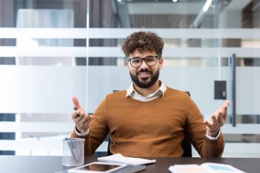 Gözlüklü ve sakallı genç bir işadamı konferans masasında oturmuş, çağdaş bir ofis ortamında kurumsal bir tartışma ya da danışma sırasında fikirlerini paylaşıyor ve iletişim kuruyor.