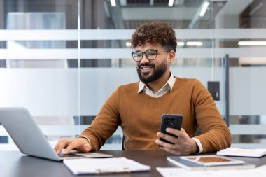 Masada oturmuş, akıllı telefonuna bakarken aynı anda bilgisayarda yazan, modern ofis ortamında verimli bir şekilde çalışan, gülümseyen sakallı genç bir adam.