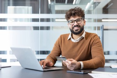 Genç adam masasında otururken kameraya gülümsüyor, bir kredi kartı tutuyor ve dizüstü bilgisayarında yazıyor, online bankacılık ve güvenli internet alımları yapıyor.