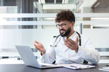 Beyaz önlüklü ve steteskoplu bir erkek doktor çevrimiçi tıbbi video konsültasyonu sırasında gülümsüyor, ofisinde bilgisayarında çalışırken konuşuyor ve el kol hareketi yapıyor.
