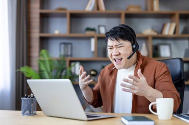 Asyalı bir adam bilgisayarındaki bir video konferansı sırasında hayal kırıklığına uğramış ve kızgın hissediyor, evde çalışırken iş stresi, patinaj ve teknik zorluklar yaşıyor