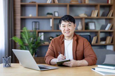 Sırıtkan Asyalı bir adam elinde küçük bir defter ve kalemle bir masada oturmuş uzaktan çalışmayı ve üretkenliği gösteriyor.