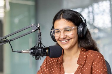 Genç bir kadın podcast yaparken gülümsüyor, kulaklık takıyor ve profesyonel bir mikrofona konuşuyor, içerik oluşturma ve iletişimi temsil ediyor.