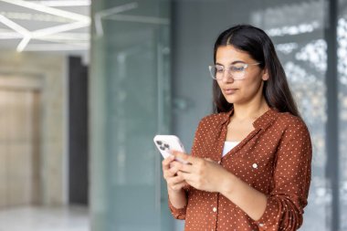 Gözlüklü ve benekli tişörtlü genç bir kadın, çağdaş bir ofis ortamında iletişim ve teknolojiyle uğraşırken cep telefonuyla daktilo çekiyor.