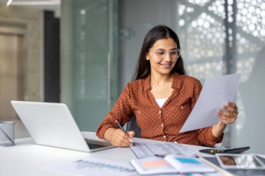 Gözlüklü genç iş kadını gülümsüyor, mali raporları inceliyor ve modern ofis çalışma alanında dizüstü bilgisayarının başında otururken dokümanlar hakkında notlar yazıyor.
