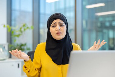 Başörtüsü takan ve sarı bluz giyen Müslüman bir iş kadını, modern ofis işyerindeki bir dizüstü bilgisayara bakarken kafası karışmış bir yüz ifadesiyle omuz silkiyor.