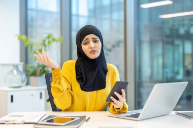 Genç Müslüman bir kadın ofis masasında oturmuş elinde akıllı bir telefonla kameraya bakarken şaşkınlığını dile getiriyor, işle ilgili sorunlar veya teknik zorluklarla boğuşuyor.