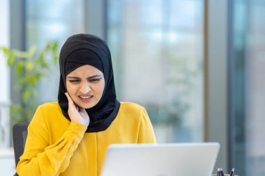 Başörtüsü takan Müslüman iş kadını, modern bir ofiste dizüstü bilgisayar üzerinde çalışırken çenesinde ve boynunda rahatsızlık ve ağrı hissederek stres veya sağlık sorunlarını işaret ediyor