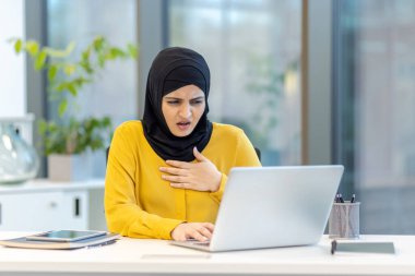 Modern bir ofiste masa başında oturan başörtüsü takan Müslüman bir kadın dizüstü bilgisayarda üzücü bilgiler okurken şok ve stres yaşıyor.