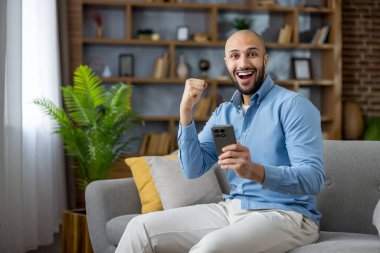 Sakallı bir adam oturma odasındaki modern bir koltukta oturuyor, gülümsüyor ve yumruğunu kaldırıyor. Akıllı telefonuyla iyi haberleri kutluyor.
