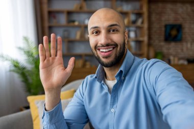 Gülümseyen ve el sallayan genç bir adam evden gelen sıradan bir video görüşmesi sırasında, dostça sanal çekim bağlantıyı gösteriyor, pozitif uzaktan çalışma etkileşimi ve gayri resmi selfie