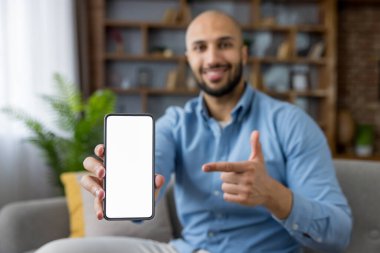 Elinde beyaz bir ekranla siyah bir akıllı telefon tutan gülümseyen genç adam, bir koltukta oturuyor ve ekranı işaret ediyor, reklam ya da başvuru promosyonu için yer sunuyor.