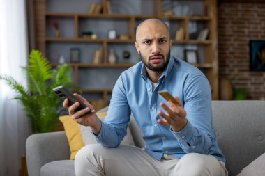 Akıllı telefon ve kredi kartı elinde tutarken kafası karışmış ve hayal kırıklığına uğramış görünen bir adam, online ödeme veya evdeki kanepesinde reddedilmiş bir işlemle ilgili sorunlar yaşıyor.