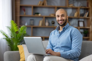 Evinden çalışan ya da ders çalışan gülümseyen genç bir adam, bir kanepede dizüstü bilgisayarla rahatlamış, uzaktan yazma, çevrimiçi öğrenme ya da modern bir oturma odasında video çağrıları yapıyor.