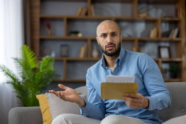 Şok olmuş genç adam evde beklenmedik bir resmi mektup okuyor, fatura ve belgelerle dolu bir zarf tutuyor, stresi, hayal kırıklığını ve endişeli, şaşkın ifadeyi gösteriyor.