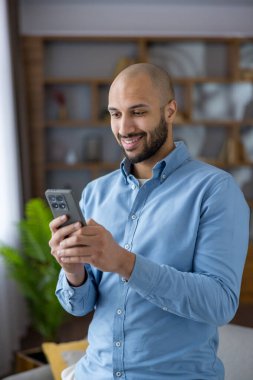 Genç adam akıllı telefonunda gülümsüyor, iletişim kuruyor ve geziniyor. Bağlantı, dijital yaşam tarzı ve çevrimiçi iletişim gibi kavramları rahat bir evde temsil ediyor.