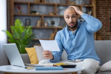 Kötü haber alan genç adam, elinde bir belge tutarken şok olmuş ve stresli görünüyor, finans yönetimi, hesaplama bütçesi ve evdeki mali baskıyla uğraşıyor.
