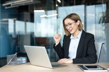 Genç iş kadını, çağdaş bir ofis ortamında dizüstü bilgisayara bakarken zaferle tezahürat yapıyor ve yumruğunu sıkıyor.