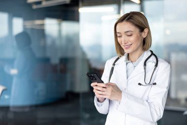 Beyaz önlüklü ve steteskoplu kadın doktor cep telefonuyla yazarken gülümsüyor, sağlık bilgilerini ve cam duvarlı hasta iletişimini yönetiyor.