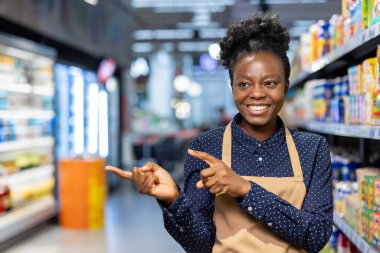 Arkadaş canlısı Afro-Amerikan süpermarket kâtibi önlüklü ve kareli gömlekli, gülümseyen ve mağaza raflarını işaret eden müşterilere yardımcı müşteri hizmetleri ve rehberlik sunan