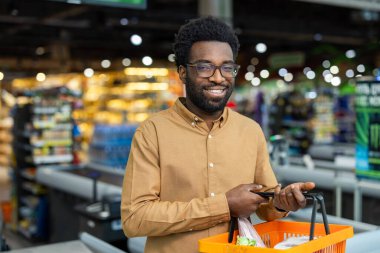 Gözlüklü ve bronz tişörtlü gülümseyen Afro-Amerikalı genç adam parlak bir süpermarket koridorunda kendine güvenen bir alışveriş sepeti tutuyor.