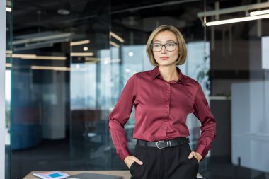 Modern bir ofiste çalışan profesyonel bir iş kadını, elleri ceplerinde, düşünceli bir şekilde kenarda duran, kurumsal bir ortamda liderliği, başarıyı ve stratejik düşünceleri somutlaştıran biri.