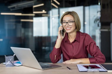 Sırıtkan iş kadını masasında cep telefonuyla konuşuyor, bilgisayar ve belgelerle iletişim ve iş görevlerini çağdaş ofis ortamında yönetiyor.