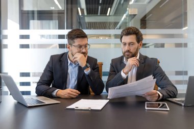 Belgeleri tartışan ve odaklanmış bir işbirliği içinde olan iki profesyonel işadamı, mali raporları gözden geçirir ve bir ofis toplantısında önemli kararlar alırlar.