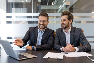 Takım elbiseli iki güler yüzlü iş adamı bir dizüstü bilgisayarın etrafında toplanarak iş stratejisi ve finansal verileri tartışıyor, ofis toplantısında takım çalışması, ortaklık ve kurumsal işbirliği sergiliyorlar.