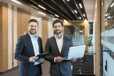Şirket koridorunda takım elbiseli iki yetişkin iş adamı. Biri bir pano, diğeri bir dizüstü bilgisayar tutuyor. İşbirliği ve profesyonel başarıyı temsil ediyor.