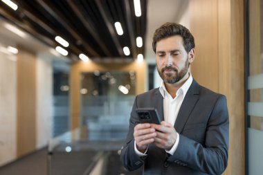Takım elbiseli bir işadamı ofis koridorunda duruyor, akıllı telefonundaki mesajları yazmaya ve kontrol etmeye odaklanmış, hareket halindeyken bağlanıyor, ağ kuruyor ve işi dijital olarak yönetiyor.