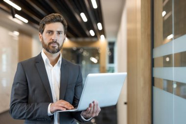 Çağdaş bir ofis binasında elinde dizüstü bilgisayarla duran profesyonel sakallı adam şirket liderliğini, yenilikleri ve kariyer başarısını temsil ediyor.