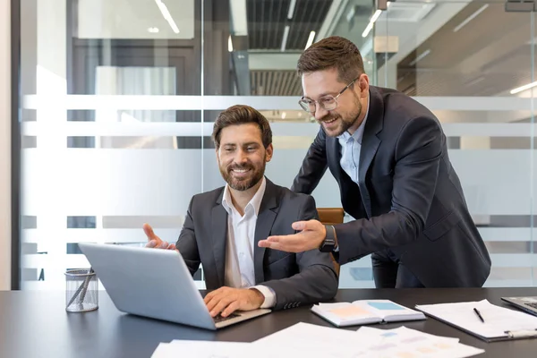 Takım elbiseli iki güler yüzlü iş adamı modern cam bir ofiste dizüstü bilgisayarda birlikte çalışıyor, üretken bir tartışmaya girişiyor ve ekranı işaret ediyorlar.