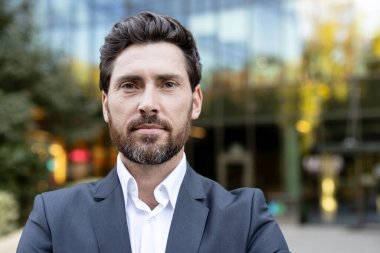 Takım elbiseli profesyonel bir adam, şehir ortamında dikiliyor arkasında bulanık bir modern bina, liderlik, başarı ve kurumsal profesyonelliği temsil ediyor.