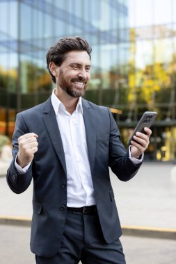 Başarılı bir iş anlaşmasını ya da başarıyı kutlayan takım elbiseli bir iş adamı elinde akıllı telefona bakıyor ve modern bir ofis binasının önünde heyecandan yumruğunu sıkıyor.