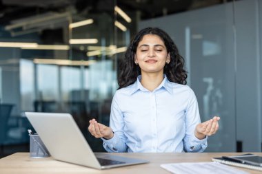 Genç iş kadını gözleri kapalı huzur ve sükunet buluyor, elleri Gyan Mudra 'da, stres yönetimi için masasında meditasyon ve farkındalık yapıyor.