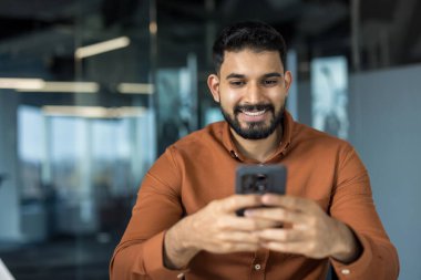 Genç adam akıllı telefonu tutarken ve kaydırırken gülümsüyor, modern ofis ortamında sosyal medya ve mobil iletişimin keyfini çıkarıyor, rahat ve kendinden emin profesyonel