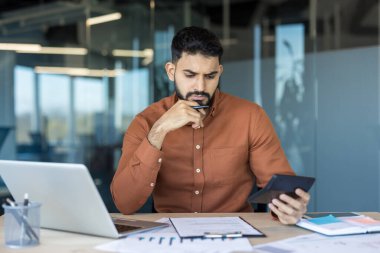 Genç Hintli işadamı mali verileri analiz ediyor ve ofisinde hesap makinesiyle hesaplamalar yapıyor, önemli bütçe ve muhasebe görevlerine odaklanıyor.