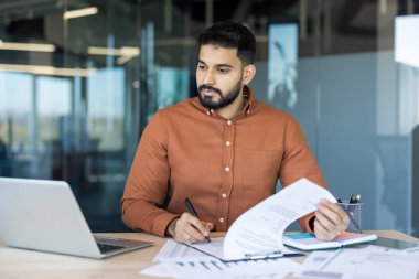 Modern ofiste çalışan genç profesyonel bir adam, evrak işlerini yönetiyor, belgeleri inceliyor ve bir dizüstü bilgisayar kullanıyor, iş süreçlerini, veri analizlerini ve şirket yönetimini temsil ediyor.