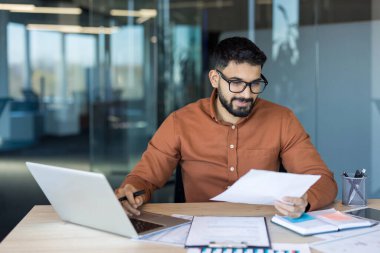 Young man in glasses and beard concentrating while working on a laptop and reviewing financial reports and paperwork, at a desk in a modern corporate office