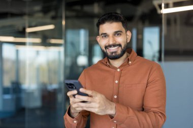 Genç Hintli işadamı, modern bir ofiste cep telefonuyla konuşurken gülümsüyor ve iletişim, teknoloji ve bağlantıyı temsil ediyor.